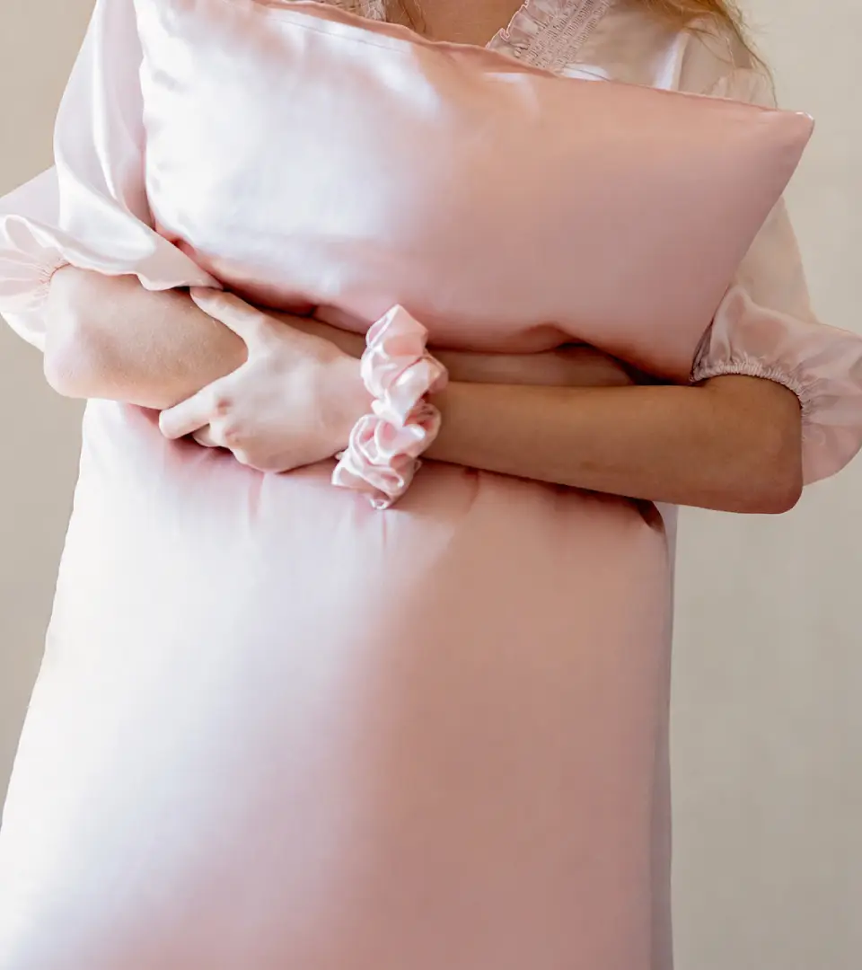 Vrouw in zijden nachtkleding met een roze zijden kussensloop in haar hand en een zijden scrunchie om haar pols
