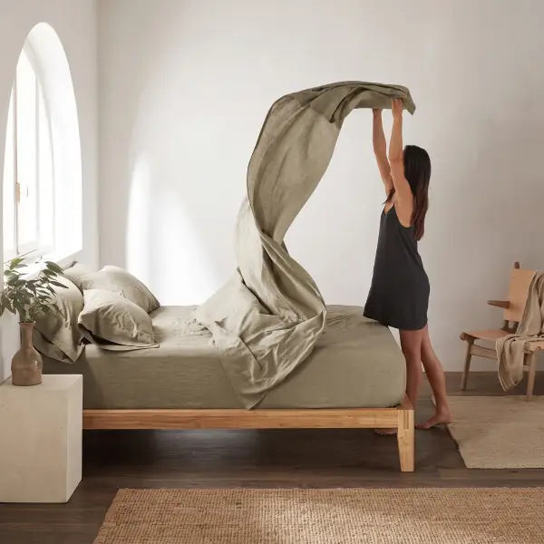 Una mujer sacude una sábana de lino verde salvia sobre una cama baja de madera en un dormitorio luminoso