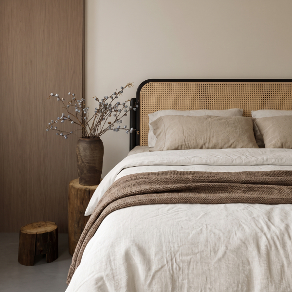 Bett in Elfenbein und Naturleinen mit Strickplaid, Kopfteil aus Rohrgeflecht und trockenen Zweigen in einer Tonvase