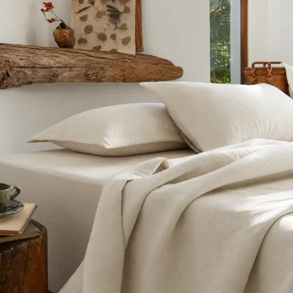 Een bed met ivoorkleurige katoenen lakens en een losjes gevouwen plaid onder een rustieke houten plank