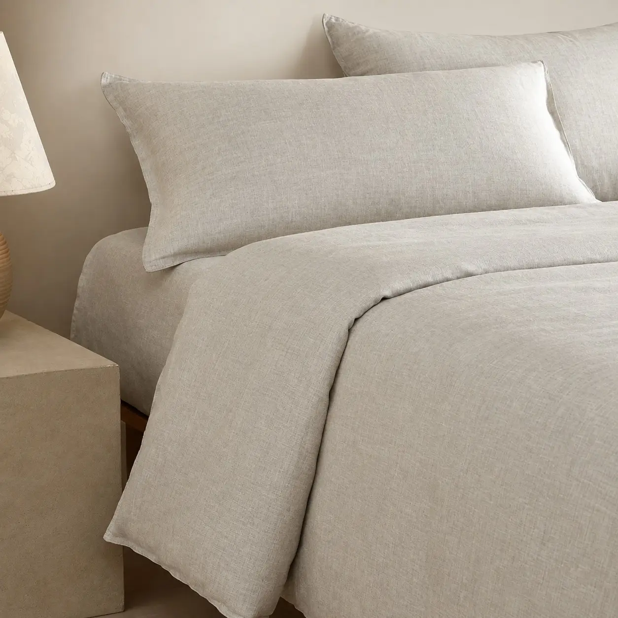 Letto con biancheria in canapa grigio chiaro accanto a un comodino in pietra e a una lampada da tavolo