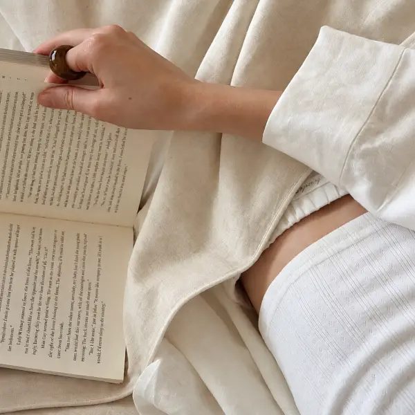 Eine Person in cremefarbener Loungewear aus Hanf liest im Bett ein aufgeschlagenes Buch