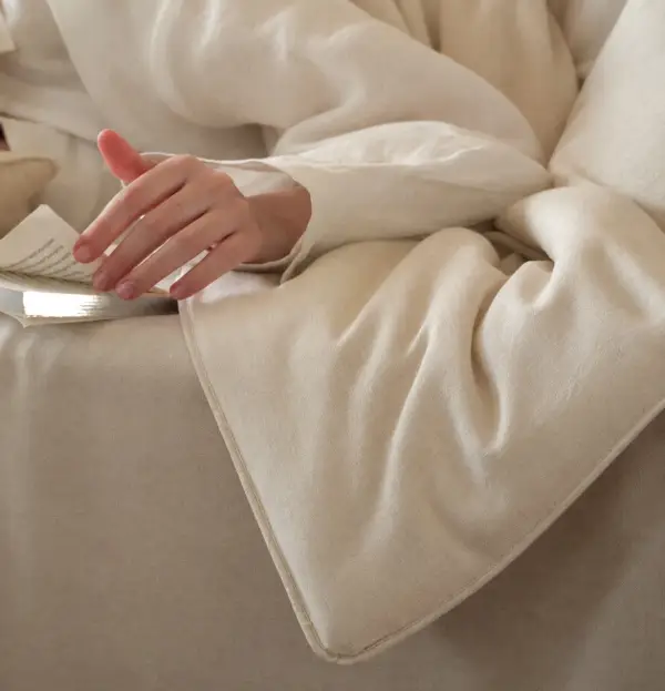 Eine Hand ruht beim Lesen im Bett auf einer weichen, cremefarbenen Bettdecke aus Hanf