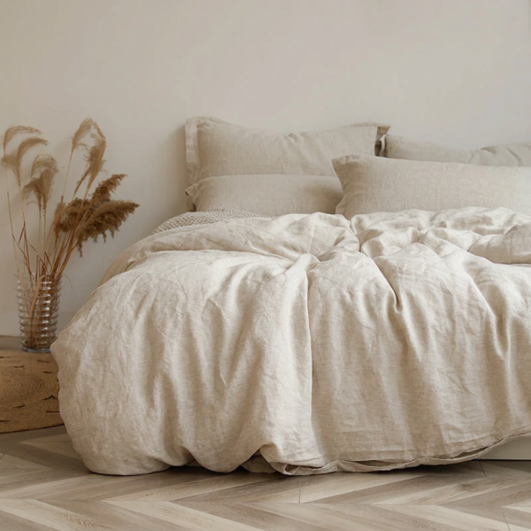 Een opgemaakt bed met ivoorkleurig gewassen linnen beddengoed en gedroogde grassen in een vaas